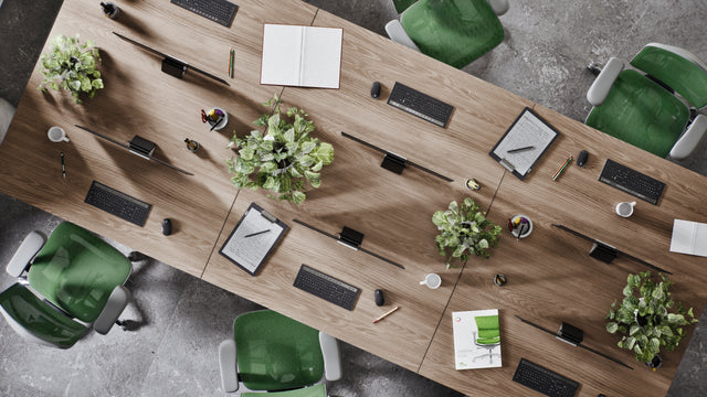 Birdeye view of a modern office space with bench desking and six monitors on each desk. At each monitor is a green mesh Enjoy office chair in a grey frame. On the desks are also clipboards, pens, and plants.