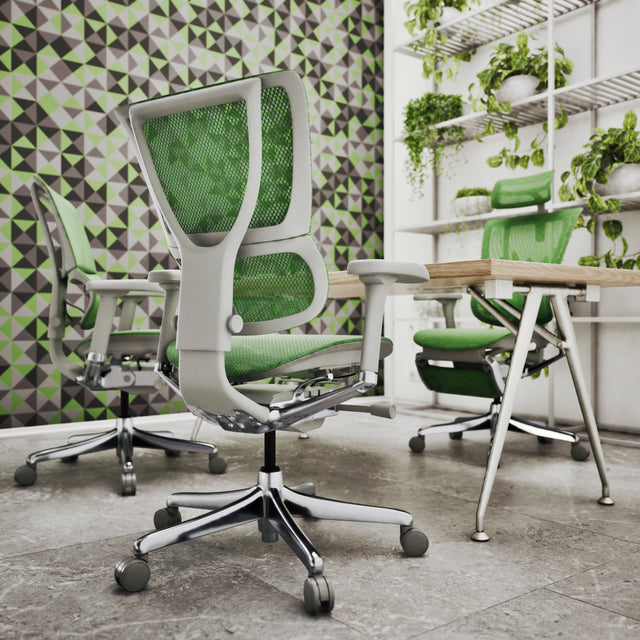 The photo is focused on the back right view of the Mirus office chair in a grey frame and green mesh. It's at a bench desk with two more ergonomic office chairs. The office has grey tiled floors and green geometric wallpaper. On the back wall is shelving with vibrant green plants.