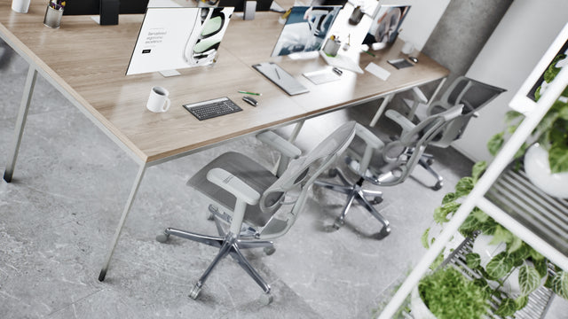 An angled view of an office bench desk with a light wood top. There are three Mirus chairs at the desk. These comfortable office chairs are in a grey frame with grey mesh. On the desks are three monitors with the ergohuman.store website on the screen.
