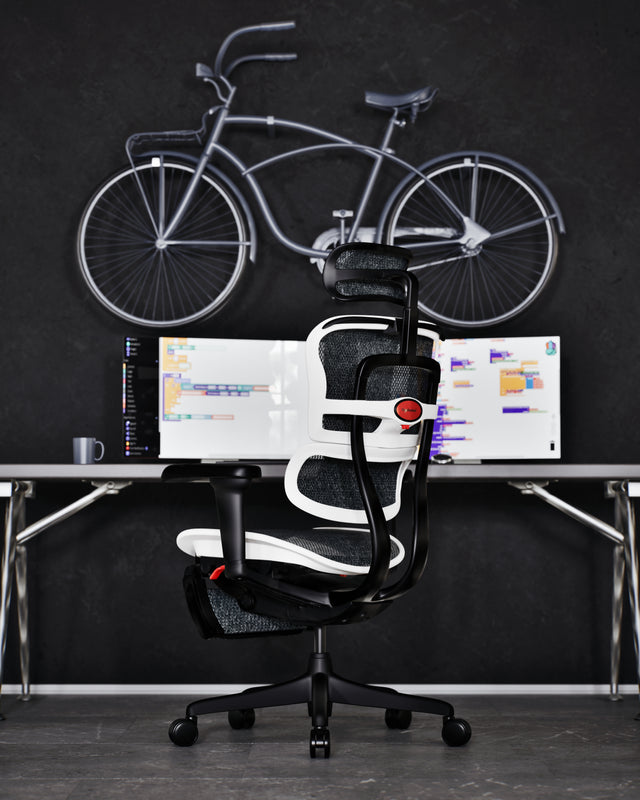 White Ergohuman Ultra gaming chair with black mesh is at a desk. On the screen is someone's work, showcasing how the gaming chair is suitable for both work and play. On the desk is a cup of coffee. Above the desk is a black bike hanging on the wall.