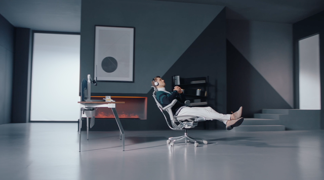 Ergohuman Elite G2 teaser video, showing a man settling into his chair. He is set in a modern, large, grey office, listening to music and drinking whisky as he relaxes. 