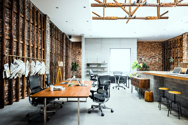 View of an office space with large bench desks and counter for coworking. The flooring is white and the walls are exposed brick. At the bench desks are black Ergohuman office chairs, great chairs for professional office spaces. 