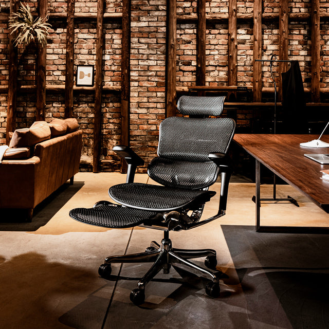 Black office chair in rustic room. The Ergohuman Elite in black frame & black mesh is reclined with the legrest unfolded. The chair is in a brown rustic room with exposed brick walls and beams. There is a brown sofa in the back left and the chair is situated at a wooden desk. 